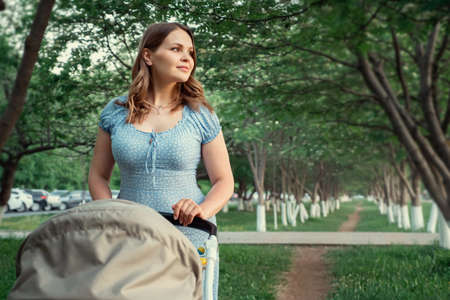 Smiling blonde lady in blue dress lulls little kid rolling grey baby carriage walking along green recreational park on sunny spring dayの写真素材