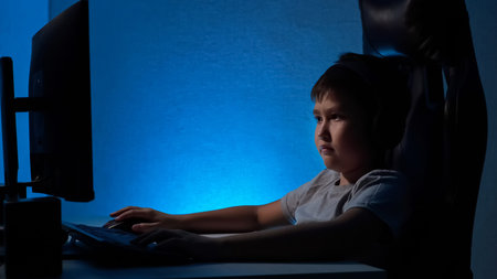 Boy with headphones playing on the computer while sitting in a dark room.