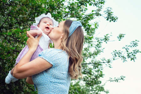 Joyful blonde mother tosses funny baby girl and kisses kid standing in local park against green trees with white blossom in spring, copyspaceの写真素材