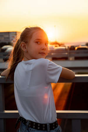 Young schoolgirl with long hair in ponytail in t-shirt looks at sunset, standing on overpass bridge backside viewの写真素材