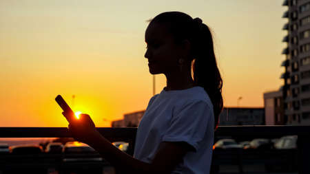 Schoolgirl looks into smartphone at sunset. Addicted young teenage girl with ponytail holds gadget in hand standing on bridge at back setting sun in summerの写真素材