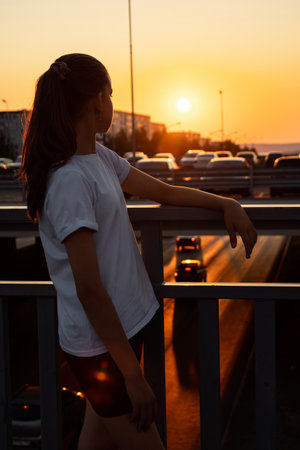 Teenage girl with long hair in ponytail in t-shirt looks at sunset, standing on overpass bridge backside viewの写真素材