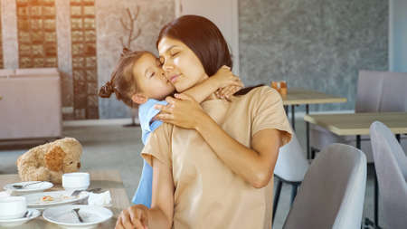 Toddler daughter hugs mom. Little child girl hugs Asian mum brunette with smile near table with white tea cups in local restaurant close viewの写真素材