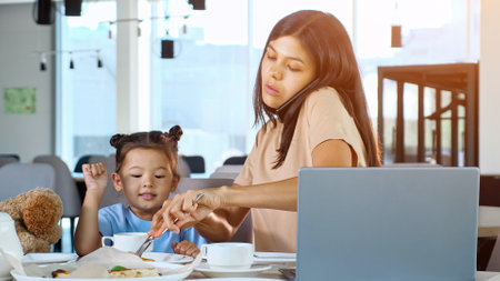Hungry Daughter asks Mother to feed himself in restaurant. Asian businesswoman mom gives pizza slice to toddler child sitting at table with tea and grey laptop close-upの写真素材