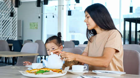Mother pours tea for girl in restaurant. Asian mom holds teapot over white cup sitting by toddler daughter and pizza on table in local cafe close viewの写真素材