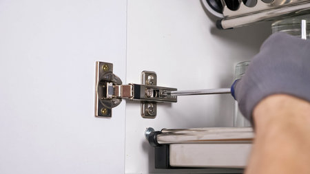 Skilled employee hands in grey textile gloves install door of white cabinet twisting screws in...