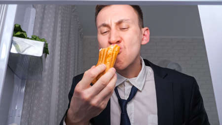 Hungry overworked businessman eats cucumber with cakes and takes plate of snacks in dark kitchen at night close view from inside refrigeratorの写真素材