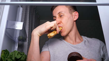 Sleepy hungry man in grey t-shirt looks for snacks and takes energy bar from shelf in dark kitchen at night, close view from inside refrigeratorの写真素材