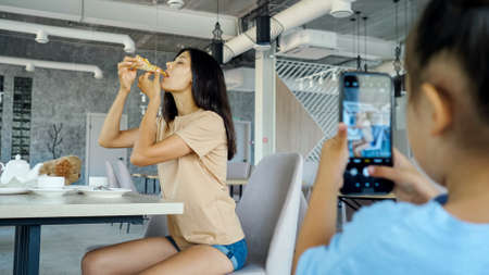 Kid girl films mom. Smart toddler girl holds black smartphone and makes photo of Asian mother brunette eating pizza in local restaurant close viewの写真素材