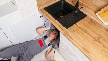 Skilled employee in grey uniform and gloves lies on floor and unscrews details repairing waste trap...