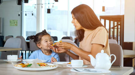 Mother feeds kid girl in restaurant. Asian brunette mom holds pizza slice and toddler daughter eats sitting at wooden table with tea in local cafe close viewの写真素材