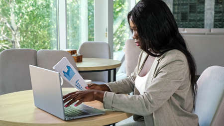 African-American lady entrepreneur types on contemporary laptop looking at chart on paper working at table in light cozy restaurantの写真素材