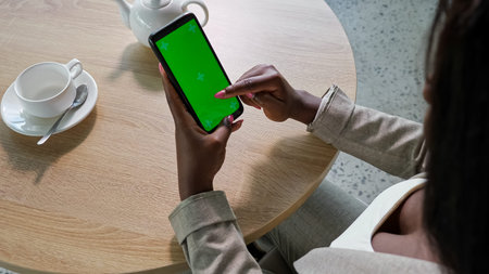 Young Afro-American woman in jacket surfs internet touching green chromakey screen of phone at table...