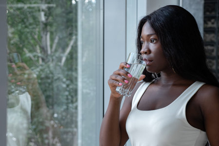 Positive young African-American woman in white sleeveless shirt drinks water looking out of large...