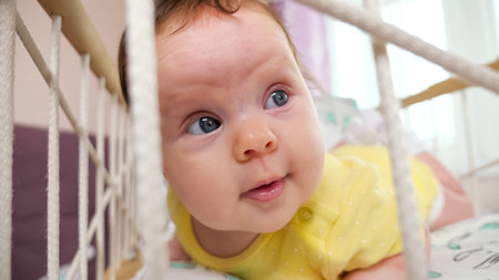 Joyful baby girl in yellow bodysuit lies on belly in rocking cradle playing and looking with...