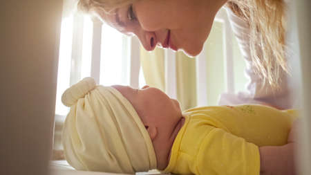 Joyful mother bends and kisses baby daughter in yellow clothing and white cap in crib against window with curtains in room extreme closeupの写真素材