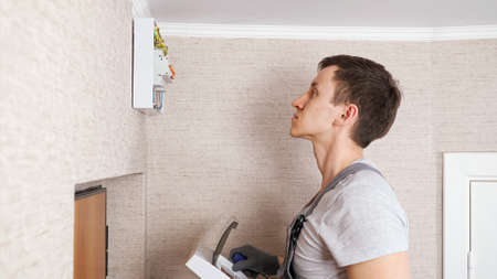 Skilled craftsman in jumpsuit covers contemporary switchboard with plastic case above front entrance door in flat hallway close side viewの写真素材