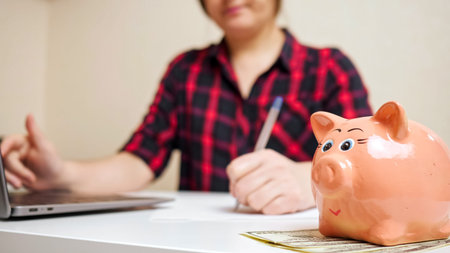 Nice pink piggy bank stands on dollar banknotes against blurred lady typing on laptop keyboard and...