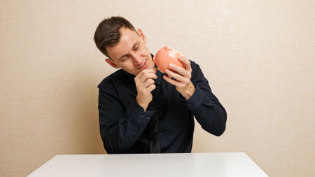 Sly man pulls dollar banknote out of piggy bank and hides bill into trendy black shirt breast-pocket sitting at white table close viewの写真素材
