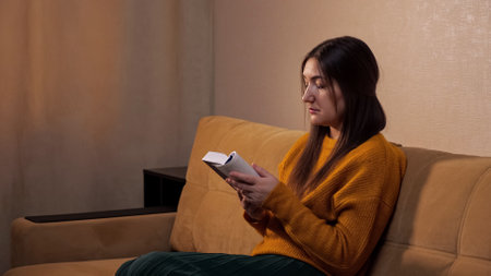 Interested lady with long loose hair holds paper book in hands and reads sitting on comfortable sofa with brown pillows slow motion closeupの写真素材