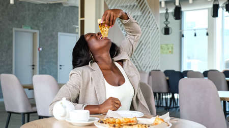 Positive African-American woman in grey jacket enjoys eating delicious pizza at table with teapot and cup in light contemporary cafeの写真素材
