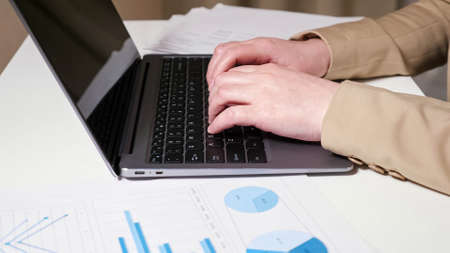 Young woman hands type on grey laptop keyboard closeup and long haired brunette freelancer looks at graphs sitting at table with coffee cupの写真素材