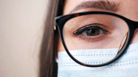 Young brunette female doctor with stylish glasses and protective mask poses for camera standing on beige background extreme cropの写真素材