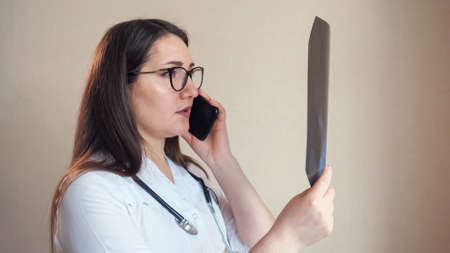 Skilled woman doctor with glasses and stethoscope talks with patient on modern mobile phone looking at X-ray picture on beige background closeupの写真素材