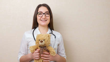 Positive brunette woman pediatrician with glasses and stethoscope holds funny teddy bear standing near beige wall in hospital closeupの写真素材