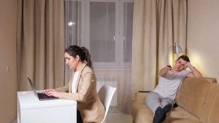 Concentrated brunette types on grey laptop sitting on chair at table and bored guy presses TV remote control buttons lying on sofa at isolationの写真素材