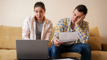 Young stressed family couple tries to calculate high payments on laptop near beige comfortable couch...