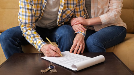 Husband signs apartment purchase contract on table near keys with house shaped breloque sitting with wife on sofa in room close viewの写真素材