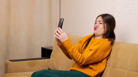 Joyful young woman blogger with long loose flowing hair sits on brown sofa and looks into black...