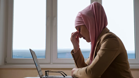 Woman in pink hijab takes a break and massages the bridge of nose while working on a laptop.