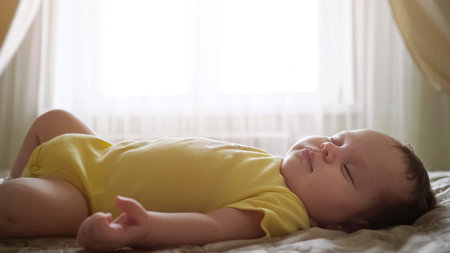 Baby girl in yellow clothing with picture sleeps in parents bed against window with transparent...