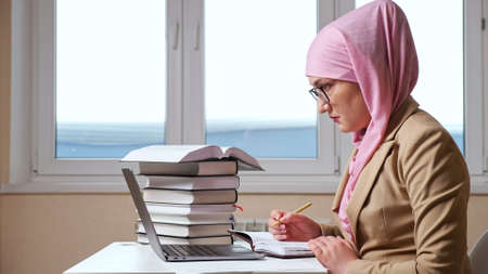Muslim woman writes with a pen in a notebook looking into a laptop.の写真素材