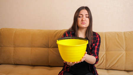 Water damage. Sad long haired woman in checkered shirt collects water flowing from ceiling into bucket sitting on sofa in living room. Concept of flooding the apartment and property insuranceの写真素材