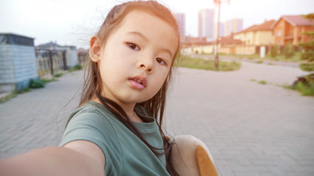 Pretty little Asian girl with soft toy dog makes selfie standing on city embankment with railing and...