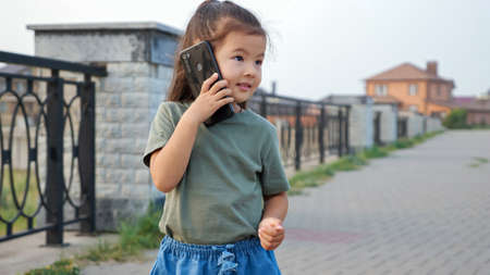 Funny little Korean girl with long ponytail talks on modern mobile phone standing on empty city embankment at sunset light close viewの写真素材