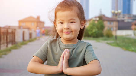 Pretty happy little Asian girl in green t-shirt holds hands in pray gesture standing on city waterfront in summer evening closeup, sunlightの写真素材