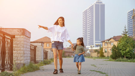 Young mother with flying hair walks with Korean little girl along contemporary city embankment pointing to beauty at sunsetの写真素材