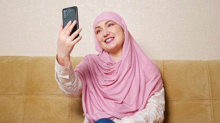 Beautiful muslim woman in hijab talking by video call while sitting on the couch.