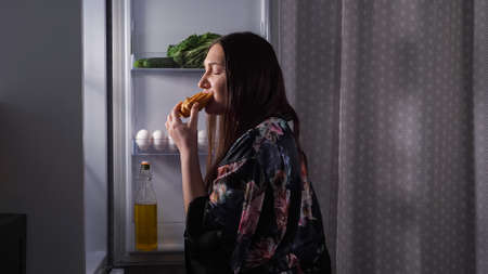 Silhouette of hungry dieting woman in elegant gown opening door of refrigerator to eat delicious eclair in dark kitchen at night close viewの写真素材