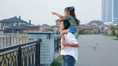 Young mommy in white t-shirt carries little Asian daughter on shoulders walking along city embankment in summer eveningの写真素材