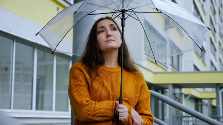 sad woman stands under a transparent umbrella in the rain on the background of an apartment building.の写真素材