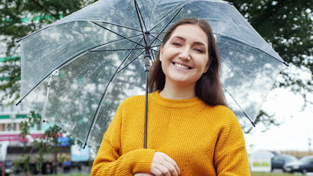 young woman in a mustard sweater stands under a transparent umbrella in rainy weather.