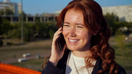 Young woman with flecks on face and copper hair talks on contemporary cellphone above street of large city at sunset light close viewの写真素材