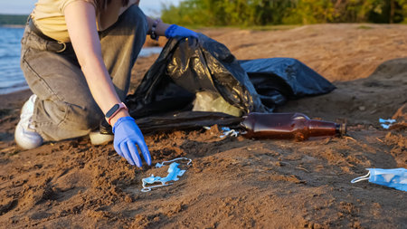 Unrecognizable woman in gloves collects trash in a black bag on the river bank, used medical masks,...