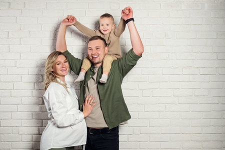Happy husband with pregnant wife and toddler boy sitting on fathers shoulders pose for photo with...