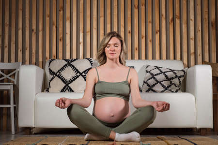 Pregnant woman with beautiful blonde curls dressed in sports set meditates sitting in lotus pose at cosy sofa on living room floor in warm atmosphere.の写真素材